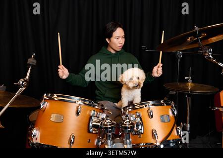 In a cozy recording studio, a young man plays the drums, sharing the moment with his dog. Stock Photo
