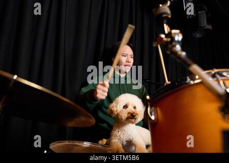 Casual young Asian man plays drums in recording studio while his adorable dog watches closely. Stock Photo