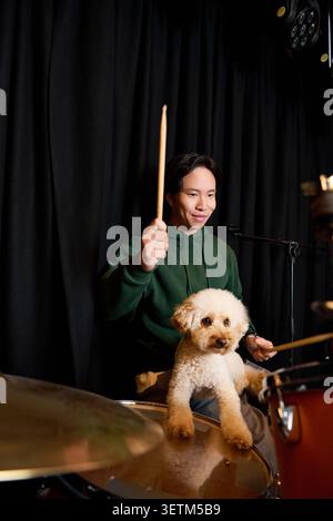 A young man joyfully plays drums while his dog sits comfortably beside him in a studio. Stock Photo