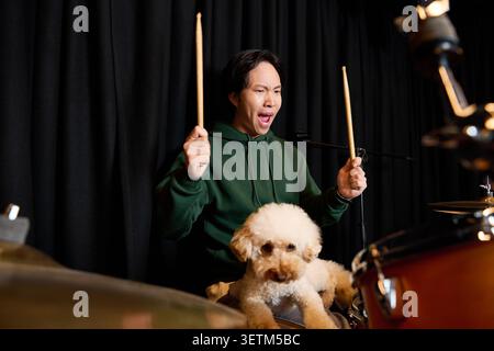 A young Asian man passionately plays drums while his playful dog sits beside him. Stock Photo