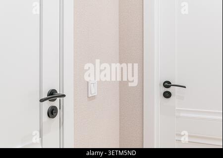 Clean interior shot showcasing two white doors with black hardware, set against a textured, light beige wall. A modern light switch is visible Stock Photo