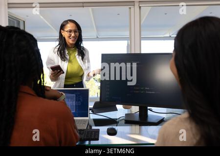 Diverse female team in office listening, leader in blazer holding phone pointing at code monitor Stock Photo