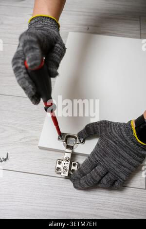 Installing concealed metal hinge on wooden panel with manual screwdriver furniture assembly process close up worker in gloves carpentry repair worksho Stock Photo