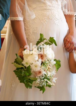 Elegant bridal bouquet of white flowers and roses held by the bride during a wedding ceremony in France, with bridesmaids in the background. Stock Photo