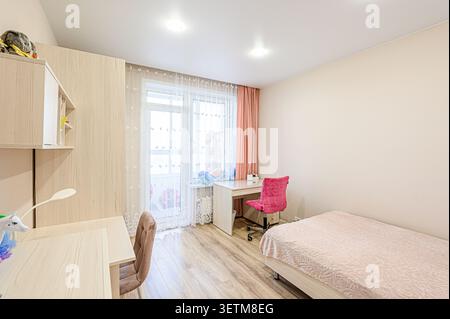 A bright, cozy room with a wooden desk, a pink chair and a bed with a patterned coverlet. Light walls and flooring give it a spacious feel Stock Photo