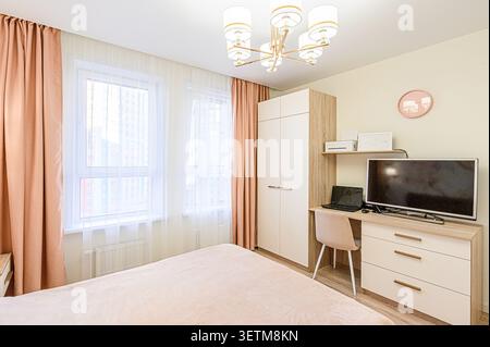 Cozy bedroom with peach curtains, large window, and a workspace featuring a desk, TV, and storage. The room has light walls and modern lighting Stock Photo