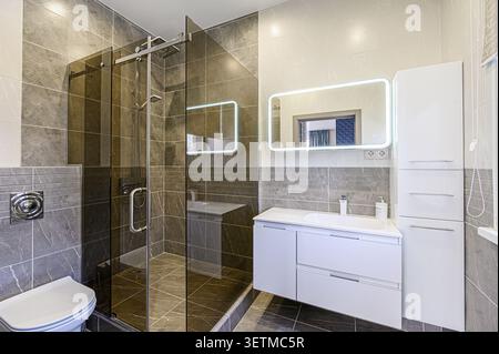 Modern bathroom with gray tile, glass shower, white vanity, and illuminated mirror. Clean, stylish design Stock Photo