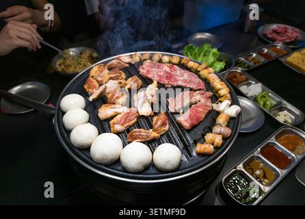 A vibrant Korean BBQ scene featuring assorted meats grilling on a circular grill. Smoke rises as diners enjoy the meal, surrounded by various side dis Stock Photo