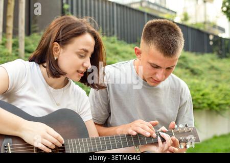 Young students and friends spend leisure time together outdoors, learning to play chords on acoustic guitar during summer vacation. Stock Photo