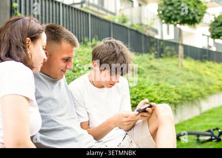 Happy diverse teenagers or students relaxing outdoors on a summer day, browsing social media and smiling while sharing content on a mobile phone. Stock Photo