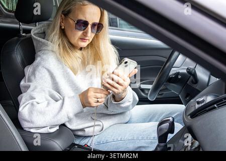 Middle aged woman sitting in parked car using smartphone connected to charging cable. Mobile communication, digital navigation preparation, travel pla Stock Photo