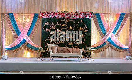 Luxury Contemporary South Asian Wedding Stage Decor with Floral Hedge Wall, Gold Geometric Frame, Chandeliers and Draped Fabrics, Empty Sweetheart Sof Stock Photo