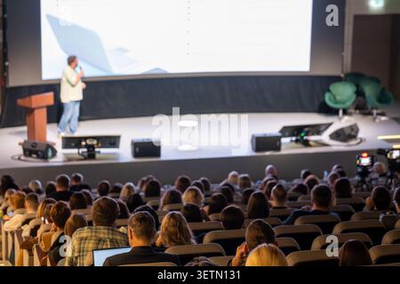 Presenter on stage delivering business presentation to large audience in modern conference auditorium, attendees listening and taking notes. Stock Photo