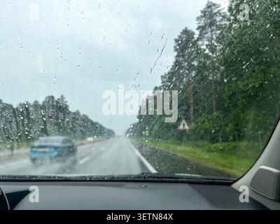 Raindrops on car windshield with blurred highway, passing cars and forest roadside, concept of bad weather driving conditions, road safety and atmosph Stock Photo