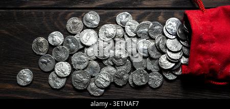 A top-down, flat lay photograph showing a collection of ancient silver coins spilling out of a red cloth pouch onto a dark, rustic wooden table. The c Stock Photo