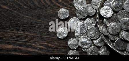A collection of ancient Roman silver denarii coins spilling from a silver bowl onto a dark, textured wooden surface. The coins feature detailed profil Stock Photo