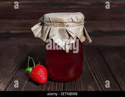 A glass jar filled with red strawberry jam, topped with craft paper and twine, standing next to a single fresh strawberry on a dark wooden table. Stock Photo