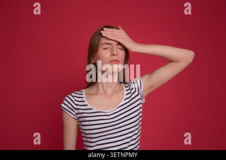 Tired young woman touching her forehead with eyes closed against a red background with copy space, headache, stress and burnout concept. Stock Photo