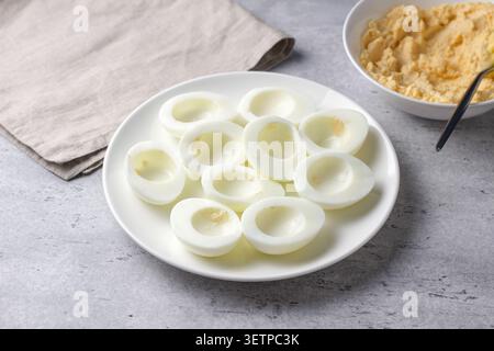 Hard boiled egg halves with yolks removed on a white plate, prepared for stuffed eggs recipe on a gray textured surface with soft natural light Stock Photo