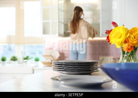 Woman putting clean dishes into cabinet in kitchen, focus on plates and vase with flowers on table Stock Photo