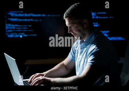 Hacker operates laptop in dark office. Hacker analyzes code screen carefully. Hacker monitors system logs for access. Hacker executes cyber attack on Stock Photo