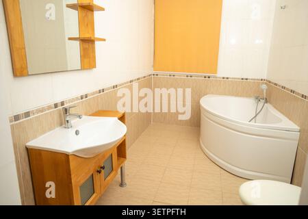 Spacious and bright modern bathroom interior featuring a white corner bathtub, wooden vanity with white ceramic sink and chrome faucet, large mirror w Stock Photo