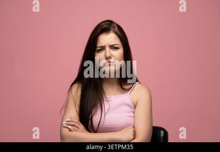 Young woman expressing skepticism and frustration, standing with crossed arms against a pink background Stock Photo