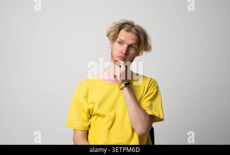 Young man with contemplative expression in yellow t-shirt pondering a problem, finding an idea, contemplating thoughts and decisions, experiencing Stock Photo
