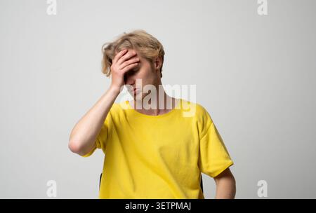 Young blond man in a yellow t-shirt suffering from intense headache or migraine, feeling stressed, tired, and frustrated while holding his forehead Stock Photo