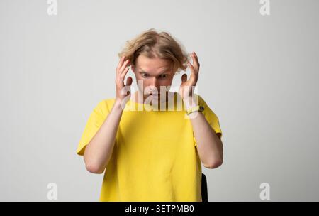 Young blond man experiencing shock and disbelief, reacting to unexpected news or a surprising event with wide eyes and hands near his head, expressing Stock Photo