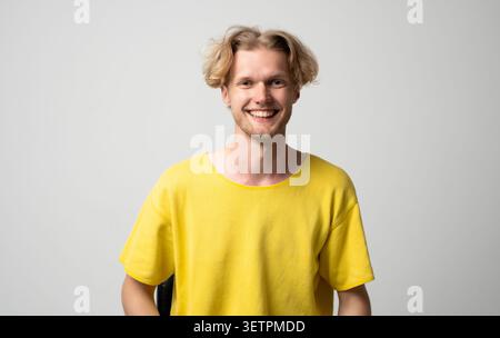 Happy young blonde man smiling confidently, radiating positivity and joy, celebrating success, feeling good, standing relaxed in a comfortable yellow Stock Photo