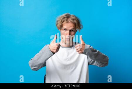 Blond young man giving double thumbs up gesture, approving excellent service, showing agreement, positivity, success, and full consent on blue Stock Photo