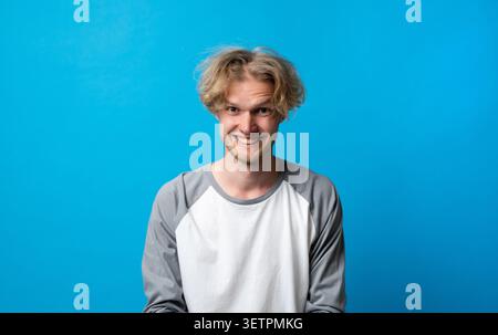 Young man with messy blond hair and a mischievous grin, posing for a cropped studio headshot against a blue backdrop, playful, confident and candid Stock Photo
