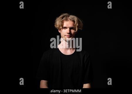 Young adult man with blonde hair looking directly at the camera, posing for a studio portrait against a dark background, representing confidence Stock Photo