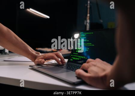 Close-up hands of unrecognizable programmer typing on a laptop keyboard displaying green code, working remotely from a home office desk, illuminated Stock Photo