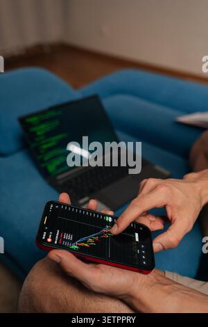 Hands holding smartphone with financial charts while laptop showing code rests on blue sofa, illustrating remote work, trading, coding, data-driven Stock Photo