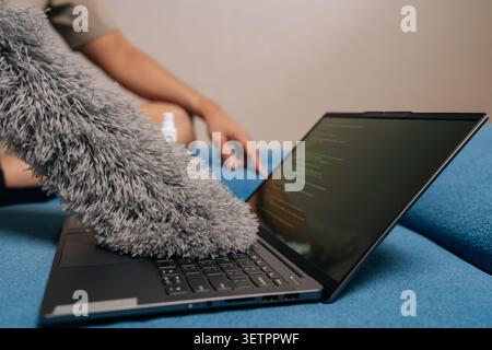 Close-up developer programmer sitting on sofa, using fluffy duster to clean up lines of messy code displayed on laptop screen, symbolizing software Stock Photo