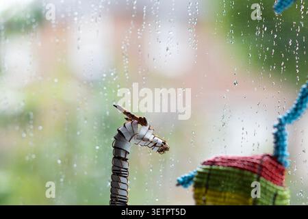 A small metal giraffe sculpture positioned beside a rain‑covered window, looking out at a cloudy, rainy day. Stock Photo