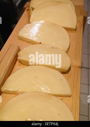 Close-up view of multiple semi hard cheese wheels cut into thick semicircular wedges arranged in row on natural wood plank board, glossy pale yellow r Stock Photo