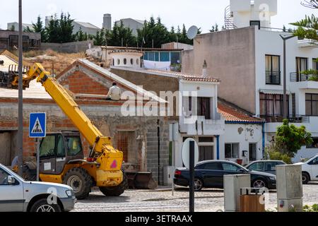 A man sits on the edge of a building under construction, with a yellow telehandler and a pedestrian crossing sign visible nearby Stock Photo