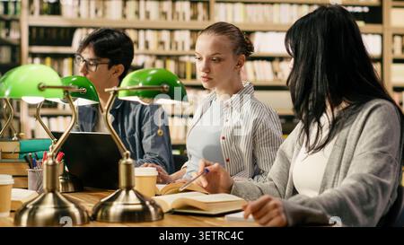University students discussing with tutor and studying with open books at library desk, taking notes and preparing for coursework. Study group using academic study materials, higher education scene. Stock Photo