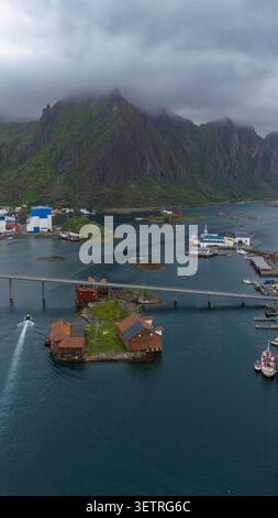 Misty dramatic peaks tower over a serene Nordic harbor with traditional red wooden cabins on stilts, fishing boats, and industrial Stock Photo
