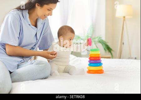 Smiling mother and baby play with colorful stacking rings on white bed at home Stock Photo