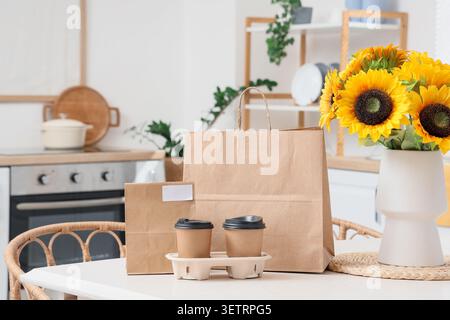Paper bags and takeaway coffee cups on kitchen table with sunflowers in vase Stock Photo