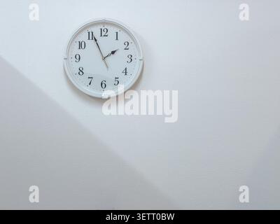 A round white wall clock showing approximately 11:10 is mounted on a smooth white wall, with a diagonal band of sunlight casting a subtle shadow below. The minimal composition offers generous copy space for text overlay. Stock Photo