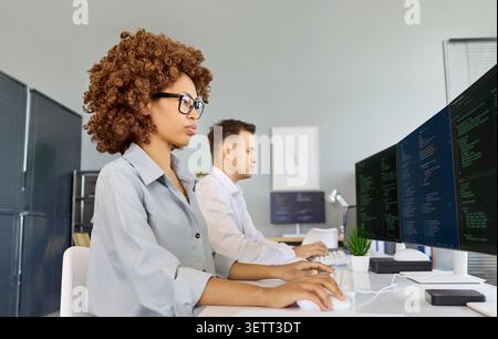 Female developer coding at multi monitor workstation with colleague in office Stock Photo