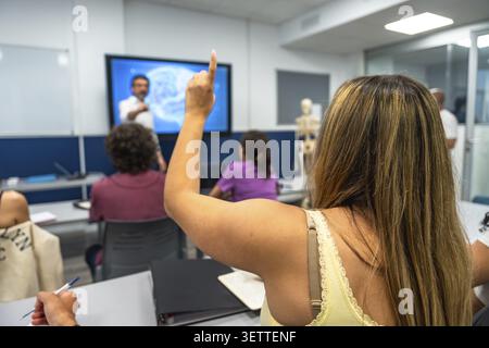 Student raising hand to ask question during a university lecture, actively participating in a learning environment with a professor teaching on an int Stock Photo