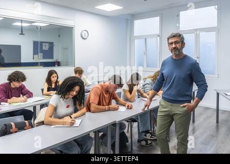 Professor standing next to desks while diverse group of college students are writing during a test, studying, or completing an assignment in a modern Stock Photo