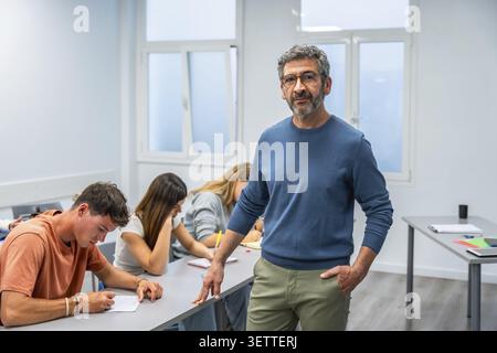 Experienced professor in smart casual attire stands in a bright modern classroom observing diverse students focused on writing during a university exa Stock Photo