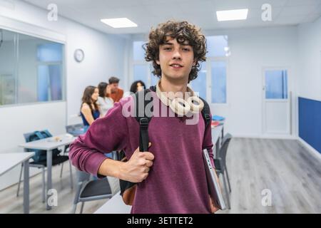 Young male student with curly hair and headphones around his neck holds a laptop and backpack, smiling confidently in a classroom while peers chat in Stock Photo
